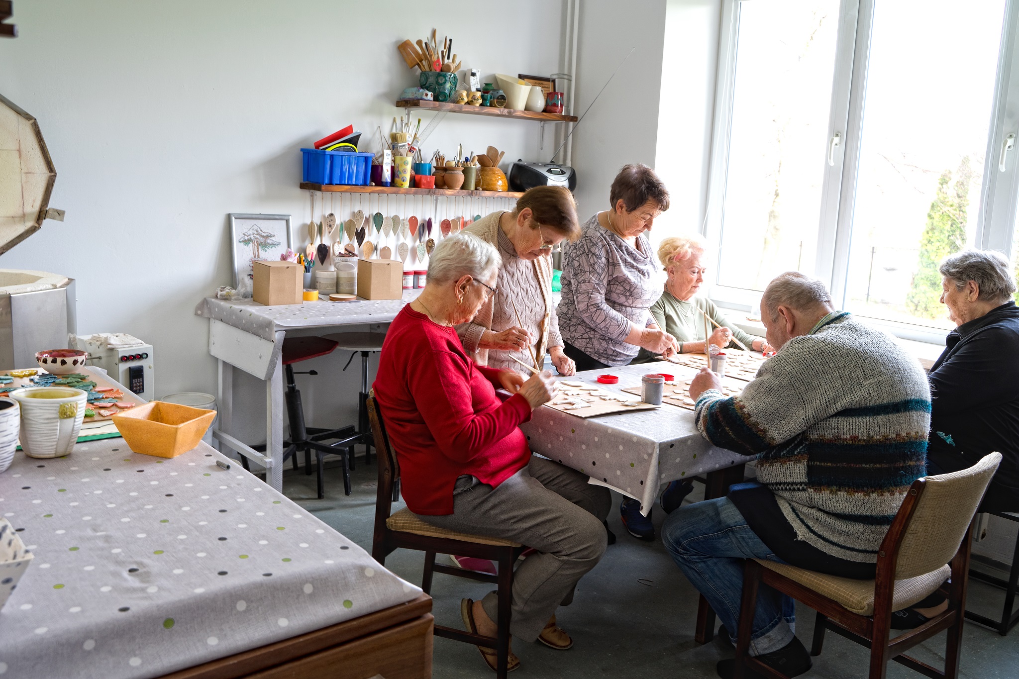 Na zdjęciu przedstawione wnętrze pracowni ceramicznej, seniorzy siedzący przy stole wykonujący ozdoby ceramiczne.