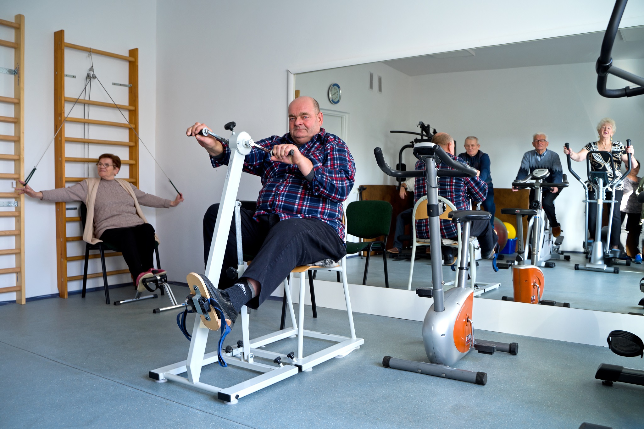 Sala zajęć ruchowych, na pierwszym planie mężczyzna ćwiczący na rowerze rehabilitacyjnym, w tle kobieta siedząca przy drabinkach w trakcie ćwiczeń rehabilitacyjnych, w lustrze na ścianie widać inne osoby ćwiczące na rowerach rehabilitacyjnych