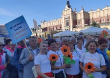 Powiększ zdjęcie dębiccy seniorzy w białych koszulkach z logo Ośrodków Wsparcia Dziennego Senior Plus zebrani trzymaja kwiaty z bibuły w dłoniach na krakowskim Rynku
